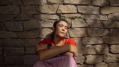 Lisa Reihana sitting with her back against a stone wall, knees drawn up and arms wrapped around each other. Her face is tilted to the right and her eyes are closed