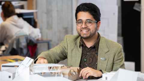 Close up of Sahil sitting at a desk with architectural models. He wears a blazer and glasses.