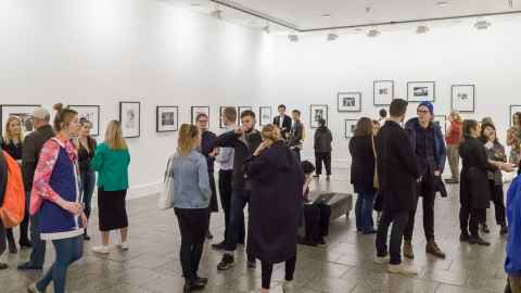 Many people in a gallery exhibition, with framed photos on the walls.